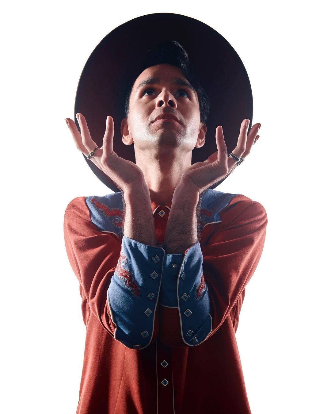 Adult male with a hat looks up with his hands raised in a studio shot. He is wearing a western-style shirt and multiple rings.